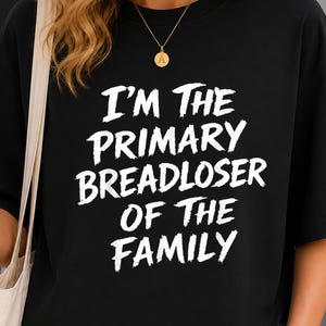 May include: A black short-sleeved t-shirt featuring white text printed on the front that reads I'M THE PRIMARY BREADLOSER OF THE FAMILY in a bold, distressed, brush-stroke style font. The shirt has a crew neckline and a relaxed, loose fit.