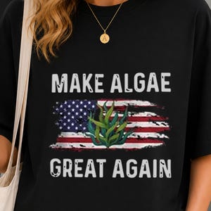 May include: A black t-shirt featuring the white text "MAKE ALGAE GREAT AGAIN" centered above and below a graphic of a distressed American flag with a green seaweed illustration overlaid in the center of the flag design.