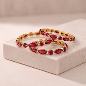 May include: Two gold rings featuring an alternating pattern of round and marquise-cut deep pink gemstones. The bands are thin and metallic, showcasing a repeating design of circular and elongated stones set securely within the gold metal frame.
