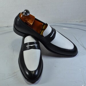 Pode incluir: Um par de mocassins penny em couro preto e branco. Os sapatos têm um design clássico com cabedal preto e gáspea e sela brancas. Os sapatos são exibidos em um fundo claro.