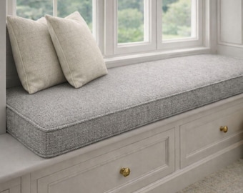 May include: A light gray rectangular window seat cushion with a textured fabric finish, accompanied by two square beige throw pillows. The cushion rests atop a built-in wooden storage bench featuring two drawers with round brass-toned knobs.