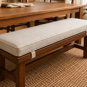 May include: A rectangular wooden bench featuring a thick, light beige upholstered cushion on top. The cushion is secured to the dark brown wooden frame with two matching fabric straps that wrap around the front edge of the bench seat.