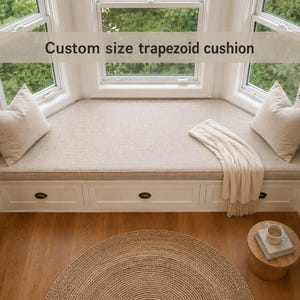 Peut inclure: Un coussin trapézoïdal sur mesure en tissu beige clair, placé sur un banc de fenêtre blanc doté de trois tiroirs. Deux coussins carrés assortis sont disposés à chaque extrémité, et un plaid en tricot crème repose sur la surface du coussin.
