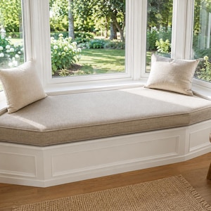 Peut inclure: Un banc de fenêtre intégré blanc doté d'un long coussin beige rembourré avec des bords passepoilés. Deux coussins rectangulaires beiges assortis sont placés à chaque extrémité du siège, installé devant un grand cadre de fenêtre.
