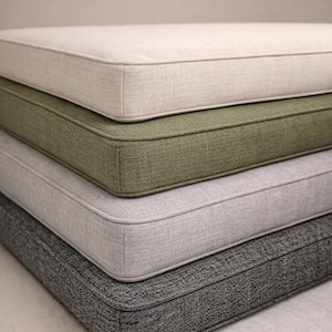 May include: Four square cushions in neutral tones: beige, olive green, light grey, and dark grey. Each cushion has a piped edge and a textured fabric, stacked to display the different colours and textures.