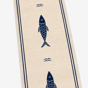 Puede incluir: Un camino de mesa rectangular de color beige con un borde de doble l&iacute;nea azul oscuro y un patr&oacute;n repetitivo de tres iconos de peces azul oscuro orientados verticalmente, intercalados con peque&ntilde;os s&iacute;mbolos de olas, con flecos en los extremos.