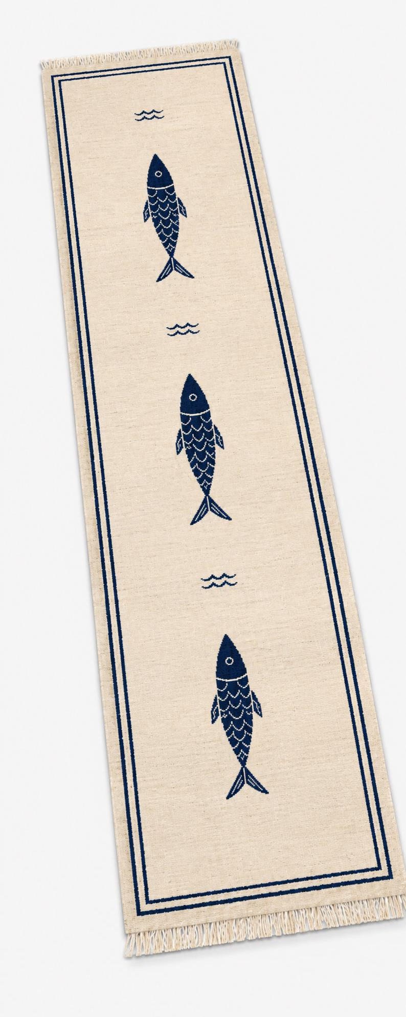 Puede incluir: Un camino de mesa rectangular de color beige con un borde de doble l&iacute;nea azul oscuro y un patr&oacute;n repetitivo de tres iconos de peces azul oscuro orientados verticalmente, intercalados con peque&ntilde;os s&iacute;mbolos de olas, con flecos en los extremos.