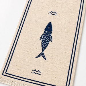 Puede incluir: Un camino de mesa rectangular de color beige con un borde azul oscuro y un patr&oacute;n repetitivo de iconos de peces y olas en azul oscuro. La tela tiene una textura tejida visible y los extremos cortos est&aacute;n rematados con flecos beige.
