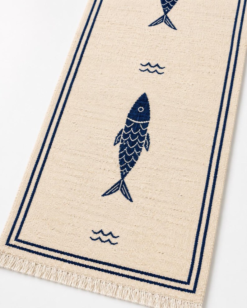 Puede incluir: Un camino de mesa rectangular de color beige con un borde azul oscuro y un patr&oacute;n repetitivo de iconos de peces y olas en azul oscuro. La tela tiene una textura tejida visible y los extremos cortos est&aacute;n rematados con flecos beige.