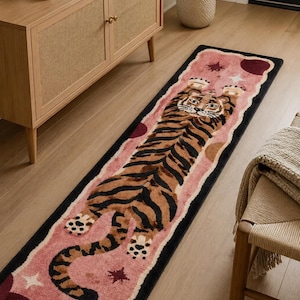 Könnte beinhalten: Ein rechteckiger Läufer mit einer zentralen Tigerillustration mit schwarzen Streifen auf orangefarbenem Fell. Der Tiger ist auf einem altrosa Hintergrund platziert, der mit dunkelroten kreisförmigen Formen und kleinen weißen Sternmotiven verziert ist, alles umrandet von einem dicken schwarzen Rahmen.