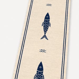 Puede incluir: Un camino de mesa rectangular de color beige claro con un borde azul oscuro y tres motivos de peces azul oscuro repetidos verticalmente. Peque&ntilde;os s&iacute;mbolos de olas azul oscuro se sit&uacute;an entre cada pez y los extremos tienen flecos.
