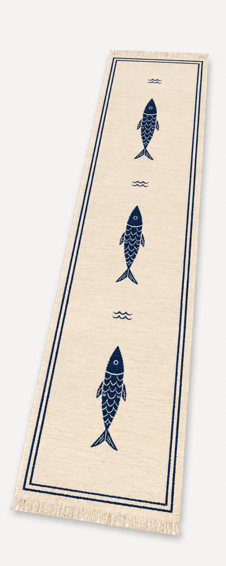 Puede incluir: Un camino de mesa rectangular de color beige claro con un borde azul oscuro y tres motivos de peces azul oscuro repetidos verticalmente. Peque&ntilde;os s&iacute;mbolos de olas azul oscuro se sit&uacute;an entre cada pez y los extremos tienen flecos.