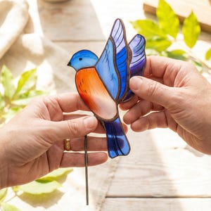 May include: A stained glass bluebird with outstretched wings, featuring shades of blue, orange, and purple. The bird has a metal stake attached for garden or yard display. The bird is held by two hands.