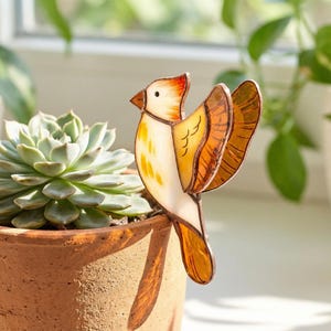 May include: A stained glass bird decoration with orange, yellow, and white hues. The bird is perched on the edge of a terracotta pot containing a succulent plant. The bird's wings are spread, and it has a detailed design.