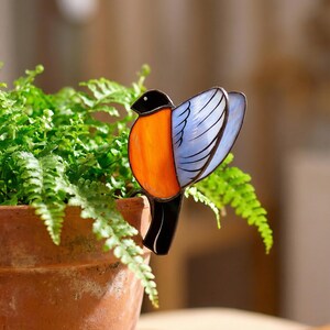 May include: A stained glass bird ornament featuring a black head, a bright orange chest, and light blue wings with dark outlines. The bird is designed to be attached to the edge of a plant pot, showcasing a vibrant color palette and glass texture.