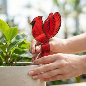 May include: A vibrant red cardinal bird garden stake with black detailing. The stained glass bird has outstretched wings and is held near a potted plant. The bird is made of red glass with black lines and a black eye.