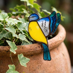 May include: A stained glass bird ornament featuring a bright yellow chest, a blue head, and wings with white and blue glass panels. The bird has dark metal soldering lines defining its shape and is perched on the rim of a terracotta pot.