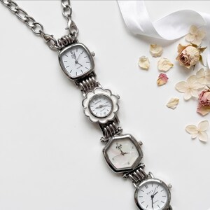 Op de afbeelding: Een zilverkleurige ketting met vier kleine horloges. De horloges hebben witte wijzerplaten en verschillende vormen, waaronder vierkant, bloem en rond. De ketting ligt op een witte ondergrond met gedroogde bloemen en een lint.