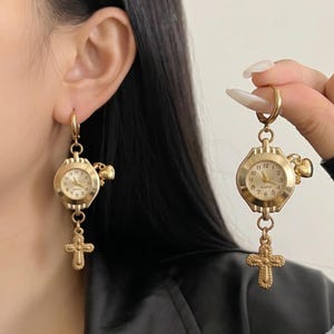 以下が含まれることがあります： 十字架ペンダント付きのゴールドトーンの時計イヤリング。イヤリングは、ローマ数字の小さな文字盤とハート型のチャームが特徴です。十字架ペンダントは時計の下にぶら下がっています。イヤリングはピアスをした耳に着用するように設計されています。