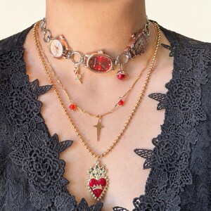 May include: A layered necklace set featuring a watch choker with red and gold accents, a gold chain with a cross pendant, and a gold chain with a red heart pendant. The jewelry is worn over a black lace top.