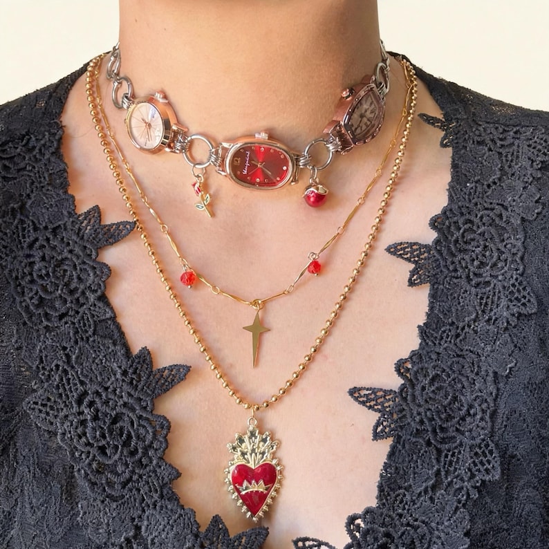 May include: A layered necklace set featuring a watch choker with red and gold accents, a gold chain with a cross pendant, and a gold chain with a red heart pendant. The jewelry is worn over a black lace top.