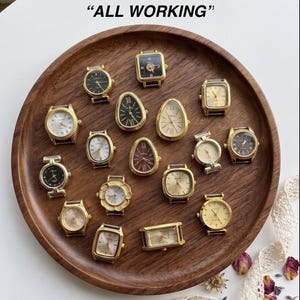 Può includere: Una collezione di vari orologi da polso color oro con diverse forme del quadrante, inclusi design rotondi, quadrati, rettangolari e a goccia. Gli orologi presentano display analogici con numeri romani o arabi su un vassoio di legno. "ALL WORKING"