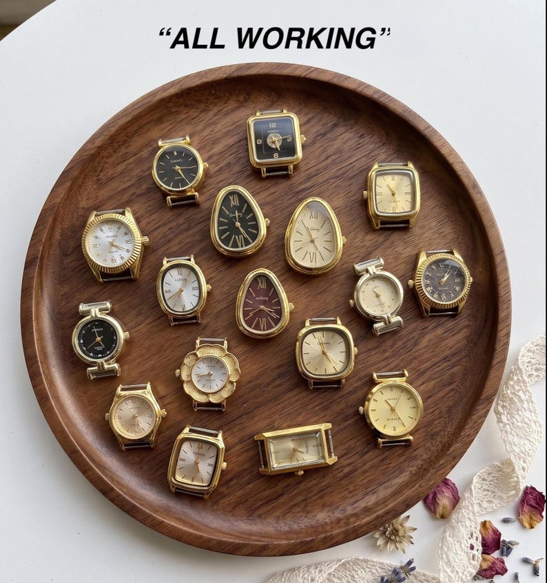 May include: A collection of various gold-toned wristwatches with diverse face shapes including round, square, rectangular, and teardrop designs. The watches feature analog displays with Roman or Arabic numerals and are arranged on a wooden tray. "ALL WORKING"