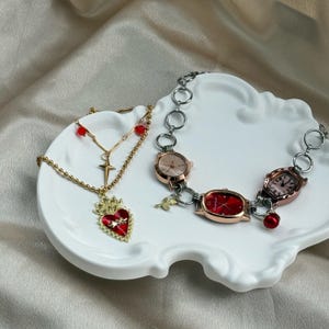 May include: A gold-toned necklace with a red heart pendant and a cross charm, alongside a watch bracelet with three watch faces. The jewelry is displayed on a white, decorative plate.