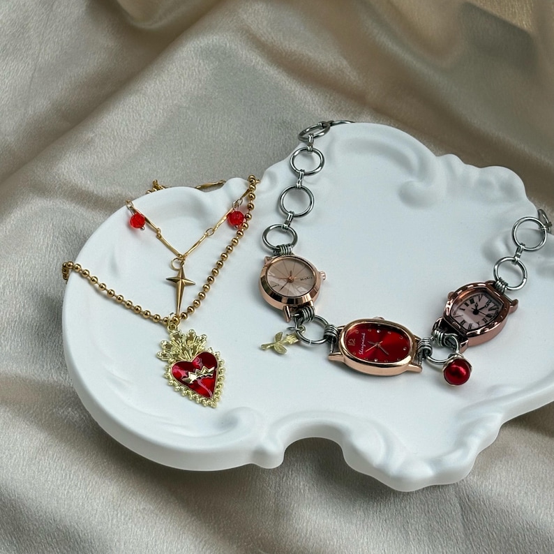 May include: A gold-toned necklace with a red heart pendant and a cross charm, alongside a watch bracelet with three watch faces. The jewelry is displayed on a white, decorative plate.