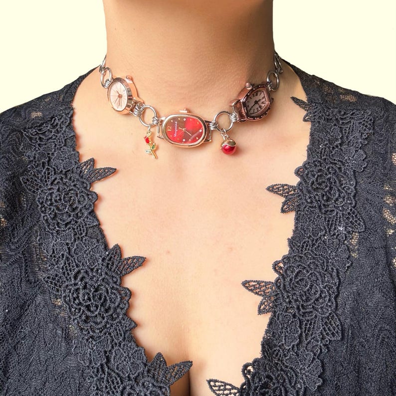 May include: A silver-toned choker necklace featuring multiple small watch faces in various styles. The necklace includes a red watch face, a rose charm, and a red bead charm. The background is a dark gray lace top.