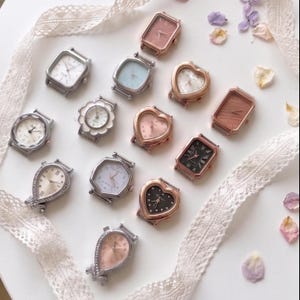 May include: A collection of twelve assorted wristwatch cases in various shapes including rectangular, heart, circular, and teardrop designs. The cases feature metallic finishes in silver, gold, and rose gold tones with diverse dial colors and markers.