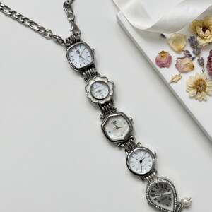 Op de afbeelding: Een zilverkleurige ketting met vijf kleine horloges met witte wijzerplaten. De horloges hebben verschillende vormen, waaronder vierkant, bloem en druppel, en zijn verbonden door een ketting. Een parel hangt aan het onderste horloge.