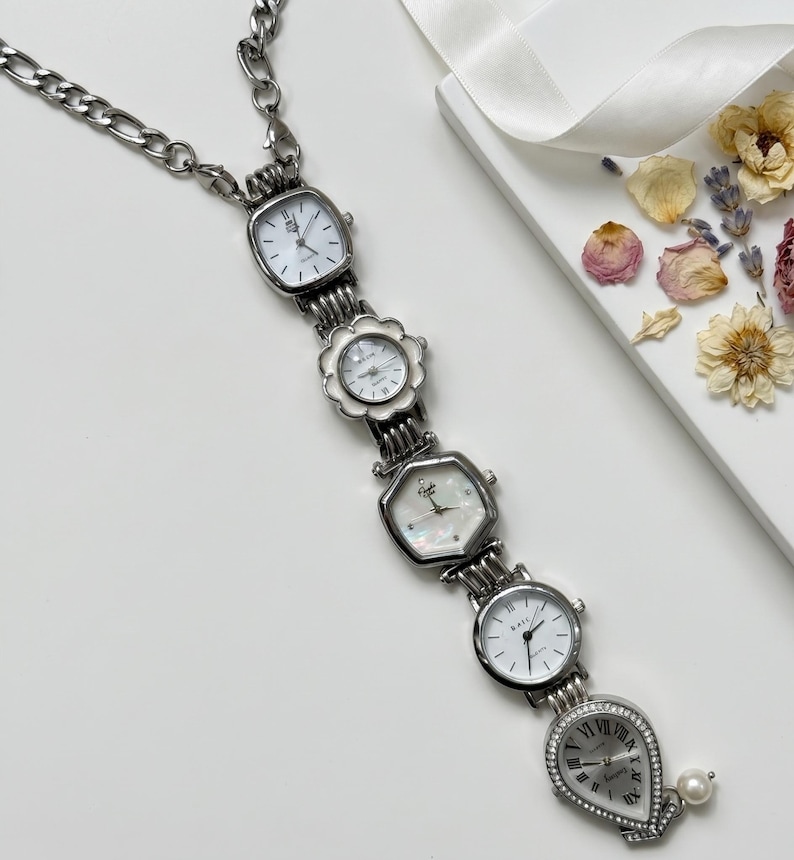 Op de afbeelding: Een zilverkleurige ketting met vijf kleine horloges met witte wijzerplaten. De horloges hebben verschillende vormen, waaronder vierkant, bloem en druppel, en zijn verbonden door een ketting. Een parel hangt aan het onderste horloge.