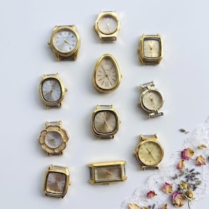 May include: A collection of eleven gold-tone watch heads in various shapes including round, square, rectangular, and teardrop. Each watch face features gold or silver dials with hour markers and hands, presented without straps on a plain white surface.