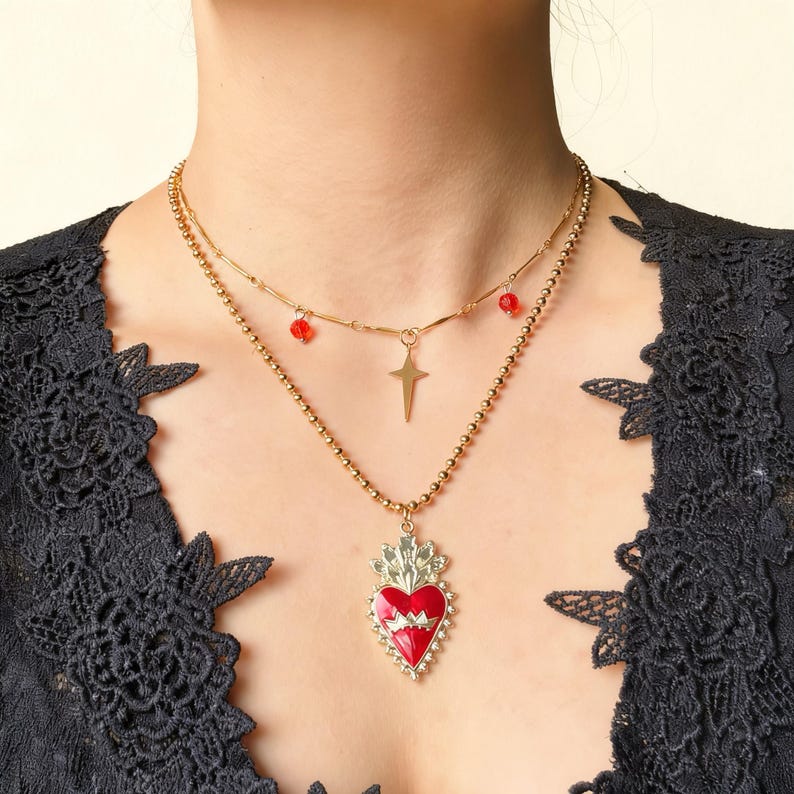 May include: A layered gold-toned necklace featuring a pendant with a red heart design, a gold cross, and red bead accents. The necklace is worn against a black lace top.