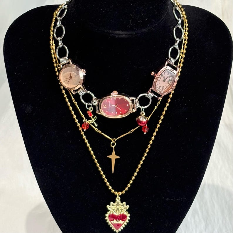May include: A multi-layered necklace featuring three watch faces with pink and red dials. The necklace includes gold and silver chains, a cross pendant, and a heart-shaped pendant with red enamel detailing. Red rose and bead accents are also present.
