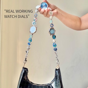 May include: A black, crocodile-textured handbag with a decorative strap made of watch dials and beads. The strap is adorned with various watch faces in shades of blue and silver, and the text "REAL WORKING WATCH DIALS" is visible.
