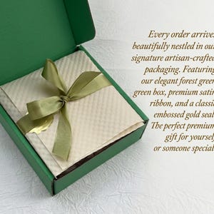 Op de afbeelding: Een bosgroene geschenkdoos met een cr&egrave;mekleurig interieur, vastgebonden met een satijnen lint. De doos bevat een cr&egrave;mekleurig item en een gouden zegel. De tekst op de doos luidt: "Every order arrives beautifully nestled in our signature artisan-crafted packaging."