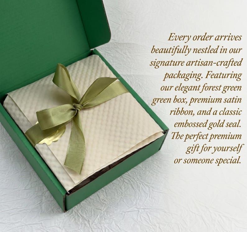Op de afbeelding: Een bosgroene geschenkdoos met een cr&egrave;mekleurig interieur, vastgebonden met een satijnen lint. De doos bevat een cr&egrave;mekleurig item en een gouden zegel. De tekst op de doos luidt: "Every order arrives beautifully nestled in our signature artisan-crafted packaging."