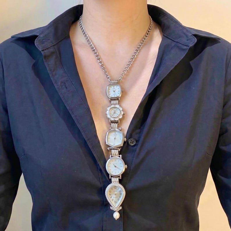 Op de afbeelding: Een zilverkleurige ketting met vijf vintage horloges van verschillende vormen en maten. De horloges zijn verbonden door een ketting en eindigen in een druppelvormig horloge met een parel. De ketting wordt gedragen over een zwart overhemd.