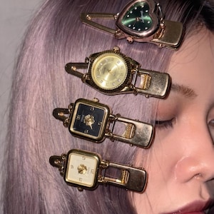 May include: Four gold-toned hair clips, each featuring a watch face. The watch faces are in various shapes: heart, round, and square. The heart-shaped watch has a green face. The round watch has a gold face. The square watches have black and white faces.