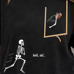 May include: A black t-shirt featuring a graphic of a skeleton running toward a football goalpost. A skeletal hand reaches out to catch a brown football near the goalpost. The text "Well, shit..." is printed in white between the skeleton and the goal.