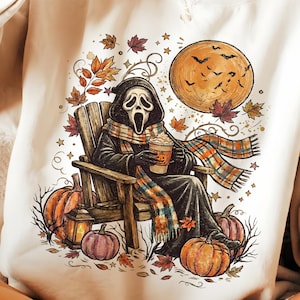 May include: A cream-colored crewneck sweatshirt featuring a graphic print of a cloaked figure with a white mask sitting in a wooden chair, holding a coffee cup, surrounded by orange pumpkins, autumn leaves, stars, and a large full moon with bats.