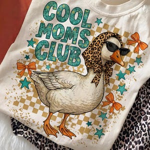 May include: Cream-colored t-shirt with the text "COOL MOMS CLUB" in teal and green. The shirt features a cartoon goose wearing sunglasses and a leopard print scarf. Orange bows and teal stars decorate the design. Leopard print pants are visible.