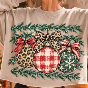 May include: A beige long-sleeve sweatshirt featuring a graphic of three Christmas ornaments hanging from green pine branches. The ornaments include a leopard print ball with a red bow, a red and white buffalo plaid ball, and a green snowflake ball.