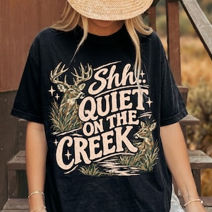 Pode incluir: Uma t-shirt preta de manga curta com uma estampa gráfica de dois veados na relva alta com o texto Shh QUIET ON THE CREEK numa fonte de estilo vintage. O design inclui pequenos detalhes de estrelas e um chapéu de palha sobre a camisa.