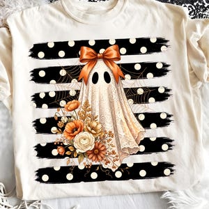 Puede incluir: Una camiseta color crema con el gráfico de un fantasma cubierto de encaje y un lazo naranja en la cabeza. El fantasma está rodeado de flores naranjas y blancas y una telaraña, sobre un fondo de rayas horizontales negras con lunares blancos.