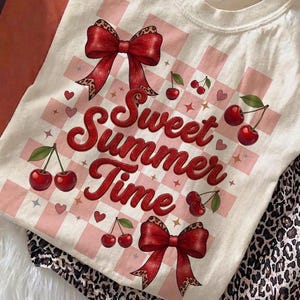 Op de afbeelding: Crèmekleurig T-shirt met een "Sweet Summer Time"-afbeelding met kersen en strikken. Het ontwerp bevat een roze geruite achtergrond, harten en sterren. Het shirt wordt gecombineerd met een broek met luipaardprint.