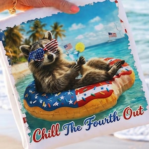 May include: White beach towel featuring a raccoon wearing American flag sunglasses and floating on a donut-shaped floatie in the ocean. The towel has the text "Chill The Fourth Out" in red and blue.