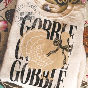 May include: A beige crewneck sweatshirt featuring a large graphic of a tan turkey with a leopard print bow around its neck. The word GOBBLE is printed in large, distressed black capital letters behind the turkey. A circular logo reads PIXEVA STUDIO.